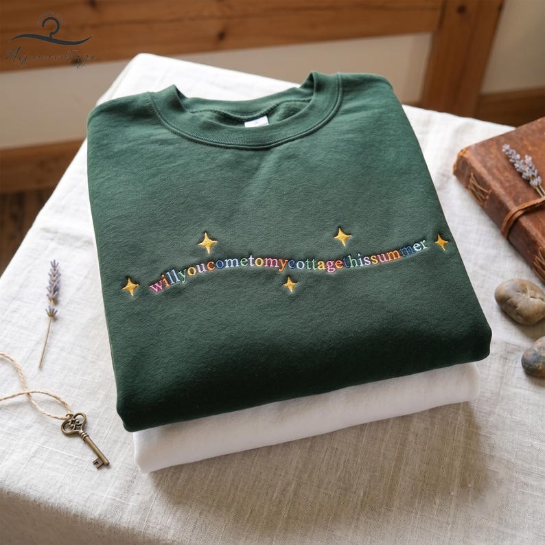 May include: A dark green sweatshirt with the embroidered text "willyoucometomycottage hissummer" in rainbow colors, accented with gold stars. The sweatshirt is folded on top of a white garment. A key and lavender sprig are visible.