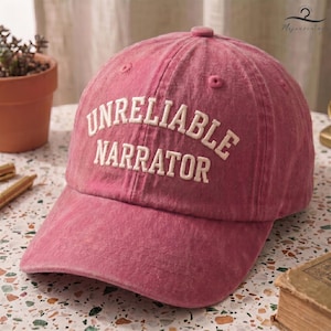 May include: A dusty rose-colored baseball cap featuring the text UNRELIABLE NARRATOR embroidered in white capital letters across the front. The cap has a six-panel construction with a button on top and a curved brim, showcasing a washed-out fabric finish.
