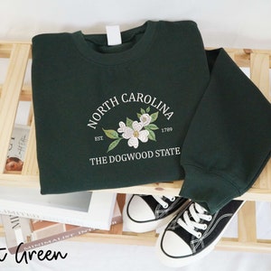 May include: Forest green sweatshirt with a white floral design and the text "North Carolina The Dogwood State". The sweatshirt is displayed on a wooden shelf with a pair of black and white sneakers.