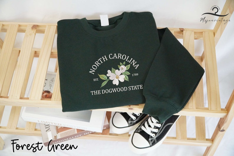 May include: Forest green sweatshirt with a white floral design and the text "North Carolina The Dogwood State". The sweatshirt is displayed on a wooden shelf with a pair of black and white sneakers.