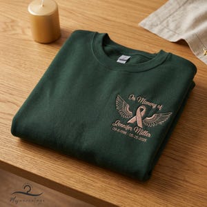 May include: Dark green sweatshirt with embroidered text and design. The design includes a ribbon, wings, and the text "In Memory of Jennifer Miller 09-11-1998 - 05-25-2025". A gold candle is in the background.