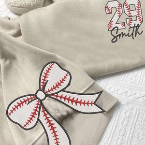 May include: A beige sweatshirt with a baseball-themed design. The sweatshirt has a large number "29" with red stitching on the front, along with the name "Smith" underneath. The left sleeve features a large red and white baseball-stitched bow.
