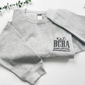 May include: A light gray sweatshirt with the text "Kat BCBA Board Certified Behavior Analyst" printed in black. The sweatshirt is folded neatly, with the sleeves and body visible. The background is white.