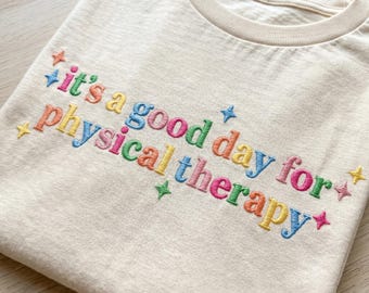 Puede incluir: Una camiseta de color crema con texto bordado multicolor que dice "it's a good day for physical therapy" en una fuente divertida. El diseño está acentuado con pequeñas estrellas bordadas de cuatro puntas en varios colores brillantes.