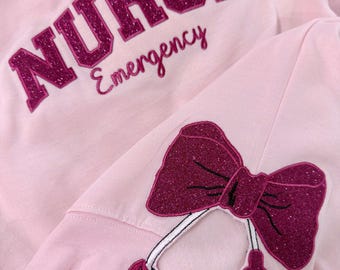 May include: Pink sweatshirt with the word "NURSE" in sparkly pink letters and the word "Emergency" written below. A sparkly pink bow with a stethoscope is on the bottom right side of the sweatshirt.