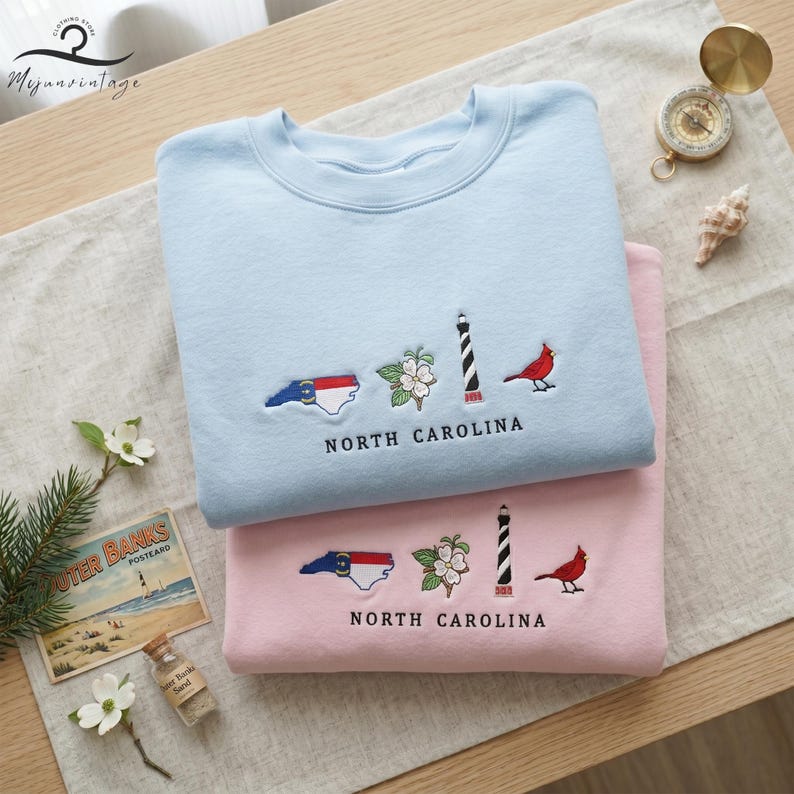 K&ouml;nnte beinhalten: Zwei Sweatshirts, eines hellblau und eines rosa, mit Stickereien aus North Carolina. Jedes zeigt die Umrisse des Staates, einen Leuchtturm, einen Kardinal und Hundebl&uuml;ten. Der Text "NORTH CAROLINA" ist darunter gestickt.
