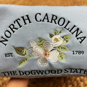 May include: Light blue sweatshirt embroidered with "NORTH CAROLINA" in a curved arch, "EST. 1789", and "THE DOGWOOD STATE". Features a floral design with white flowers and green leaves. A state-themed apparel item.