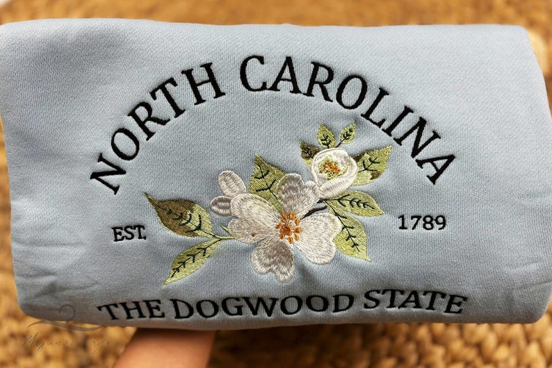 May include: Light blue sweatshirt embroidered with "NORTH CAROLINA" in a curved arch, "EST. 1789", and "THE DOGWOOD STATE". Features a floral design with white flowers and green leaves. A state-themed apparel item.