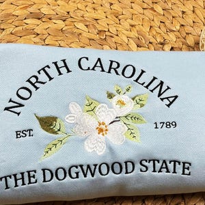 May include: A light blue fabric item embroidered with "NORTH CAROLINA" in a curved arch, a floral design, and "THE DOGWOOD STATE." The year "1789" and "EST" are also embroidered. The item is resting on a woven surface.