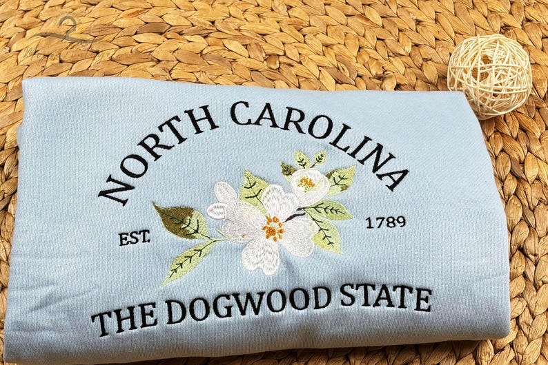 May include: A light blue fabric item embroidered with "NORTH CAROLINA" in a curved arch, a floral design, and "THE DOGWOOD STATE." The year "1789" and "EST" are also embroidered. The item is resting on a woven surface.