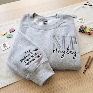 May include: A heather gray sweatshirt with the embroidered letters "SLP Hayley" on the front. The left sleeve has the embroidered text "It's a good day to help tiny humans communicate". Wooden blocks and speech bubble cards are also visible.