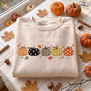 May include: A light beige crewneck sweatshirt featuring an embroidered row of five pumpkins across the chest. The pumpkins display varied patterns including polka dots, solid black with white flowers, green leaves, leopard print, and orange with white flowers.