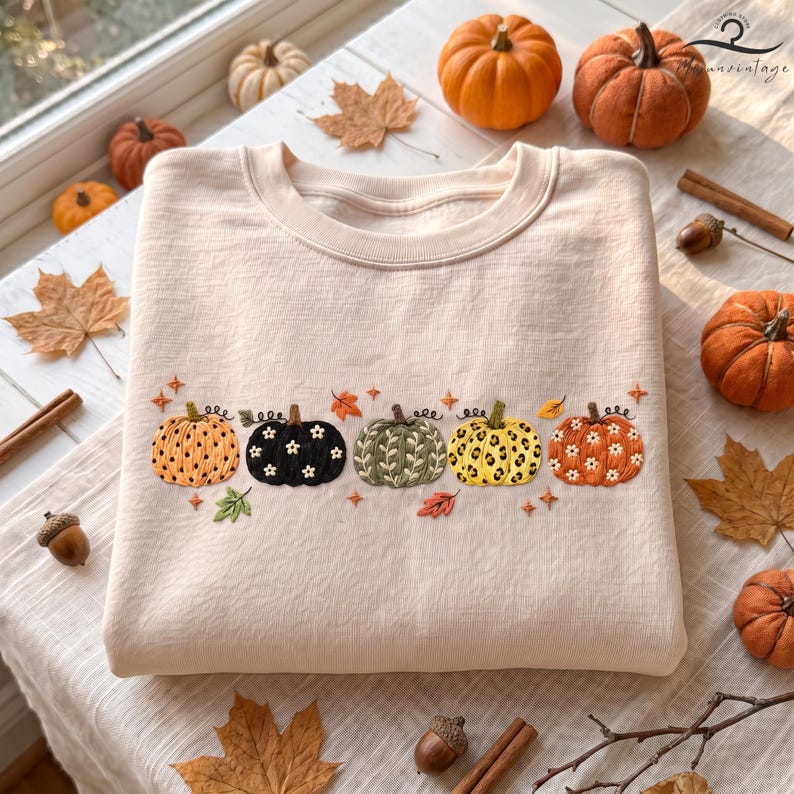 May include: A light beige crewneck sweatshirt featuring an embroidered row of five pumpkins across the chest. The pumpkins display varied patterns including polka dots, solid black with white flowers, green leaves, leopard print, and orange with white flowers.