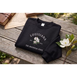May include: Black sweatshirt embroidered with "LOUISIANA", a floral design, and "THE MAGNOLIA STATE". The garment is displayed on a wooden surface, with a book and map visible in the background. The design features white and green embroidery.