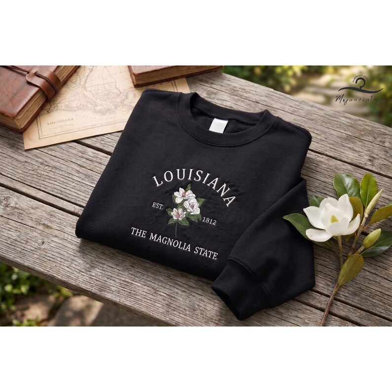 May include: Black sweatshirt embroidered with "LOUISIANA", a floral design, and "THE MAGNOLIA STATE". The garment is displayed on a wooden surface, with a book and map visible in the background. The design features white and green embroidery.
