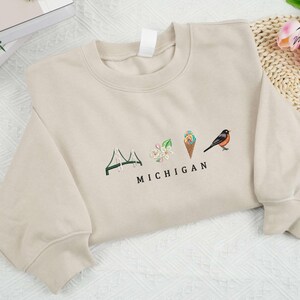 May include: A beige sweatshirt featuring a graphic design of a bridge, flowers, an ice cream cone, and a bird with the text "MICHIGAN" underneath.
