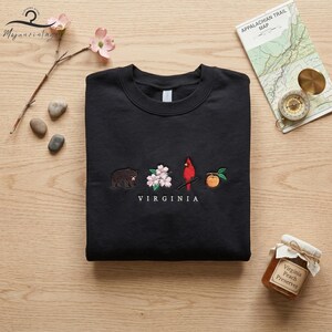 May include: Black crew neck sweatshirt featuring embroidered Virginia state symbols: a bear, dogwood flowers, a cardinal, and a peach. The word "VIRGINIA" is embroidered below the symbols. The sweatshirt is on a wooden surface.
