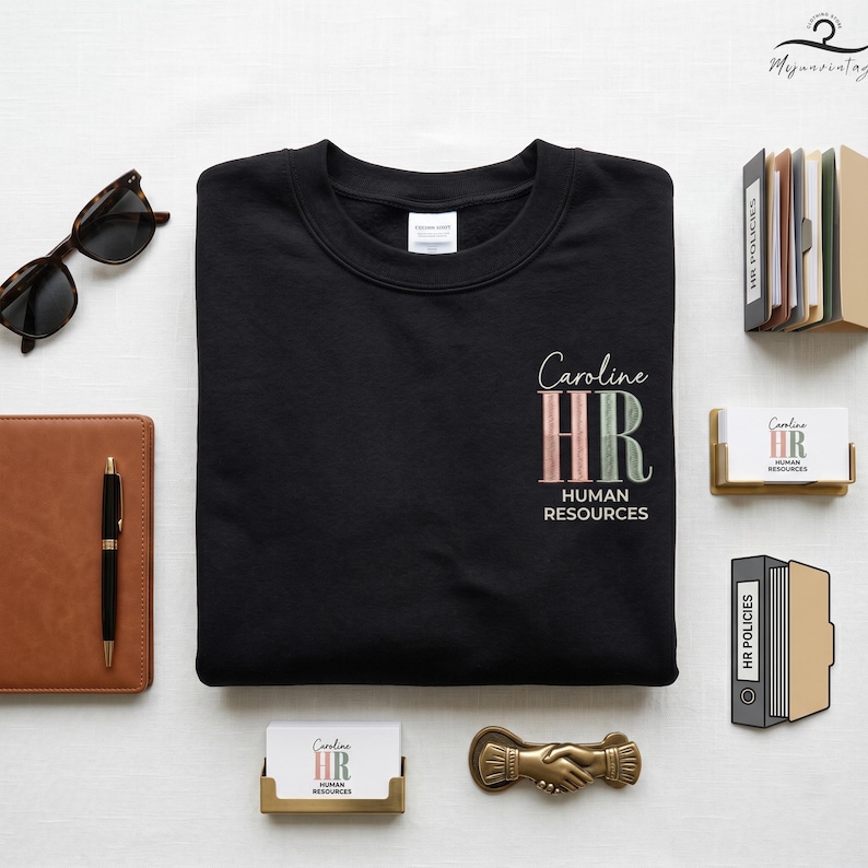 May include: Black crewneck sweatshirt with the text "Caroline HR Human Resources" in a stylish font. The design features the letters "HR" in a gradient of pink and green. Accessories include sunglasses, a notebook, and business cards.