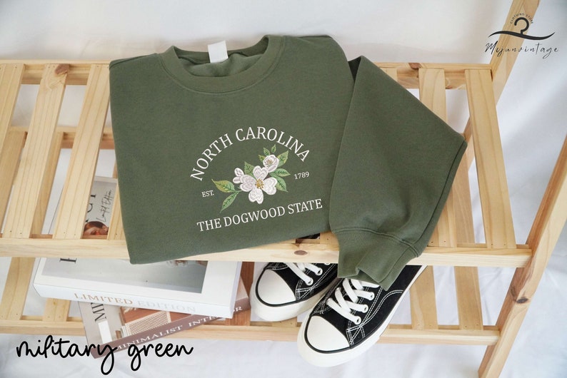 May include: Military green sweatshirt with the text "NORTH CAROLINA" arched above a floral design and "THE DOGWOOD STATE" below. The sweatshirt is displayed with black and white sneakers on a wooden shelf.