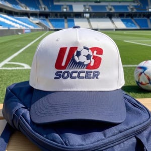 May include: A white baseball cap with a navy blue brim featuring an embroidered logo on the front. The logo displays the red letters US with a soccer ball graphic in the centre and the word SOCCER in navy blue text underneath the graphic.