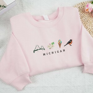 May include: Pink sweatshirt featuring a graphic design of a bridge, a flower, an ice cream cone, and a bird with the text "MICHIGAN" underneath.