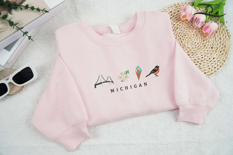 May include: Pink sweatshirt featuring a graphic design of a bridge, a flower, an ice cream cone, and a bird with the text "MICHIGAN" underneath.