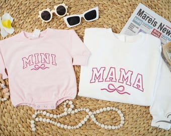 May include: Pink and white long-sleeved shirts with the words "MINI" and "MAMA" in pink lettering. The shirts are on a woven straw surface.