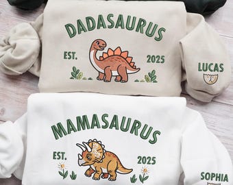 May include: Two sweatshirts, one beige and one white, with dinosaur graphics. The beige sweatshirt says "DADASAURUS" with a cartoon dinosaur and the name "LUCAS" on the sleeve. The white sweatshirt says "MAMASAURUS" with a different dinosaur and the name "SOPHIA" on the sleeve.