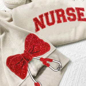 May include: A beige sweatshirt with the word "NURSE" in red glitter lettering. The sweatshirt has a red glitter bow and a stethoscope on the sleeve.