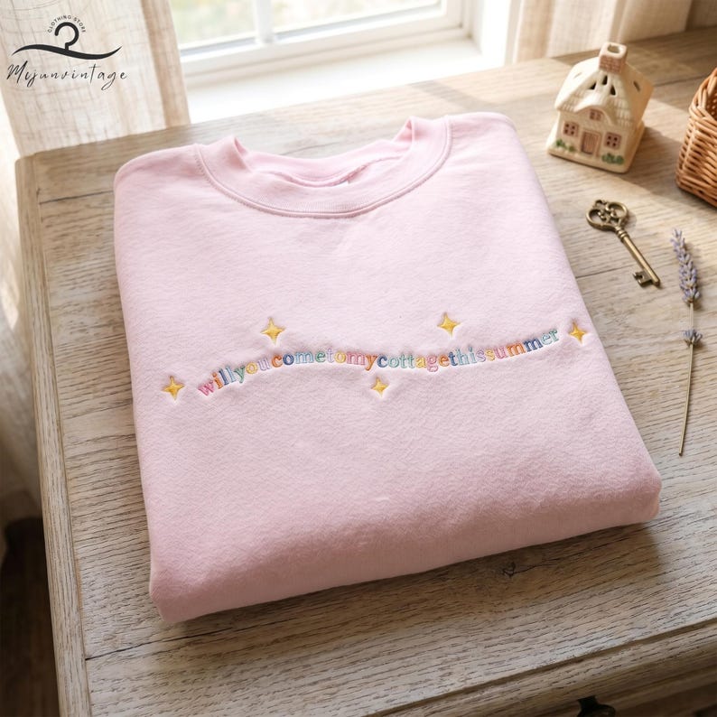 May include: A light pink sweatshirt with the embroidered text "will you come to my cottage this summer" in rainbow colors. The text is surrounded by yellow starbursts. The sweatshirt is folded on a wooden surface.