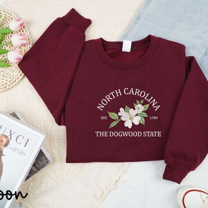 May include: Maroon sweatshirt with the text "North Carolina" and a floral design. The sweatshirt also includes the text "Est. 1789" and "The Dogwood State". The sweatshirt is displayed with jeans and white sneakers.