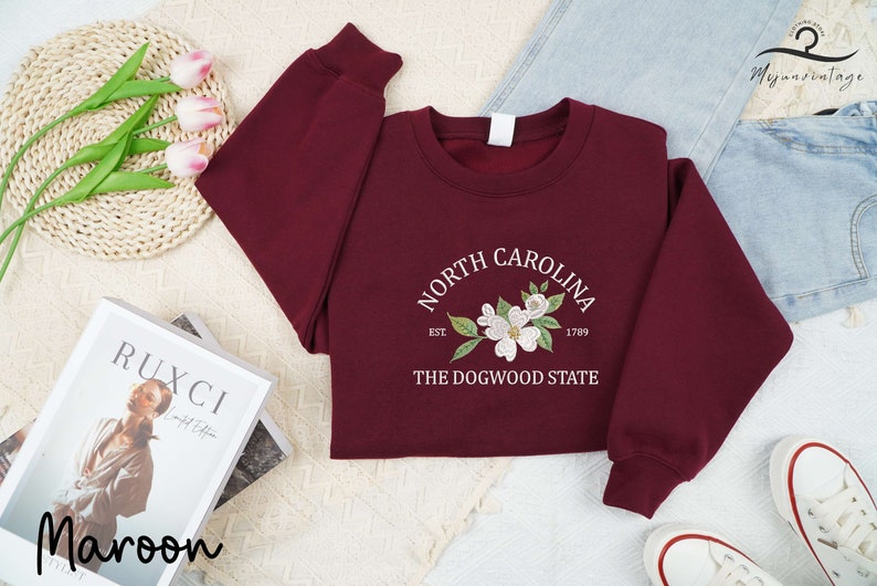 May include: Maroon sweatshirt with the text "North Carolina" and a floral design. The sweatshirt also includes the text "Est. 1789" and "The Dogwood State". The sweatshirt is displayed with jeans and white sneakers.