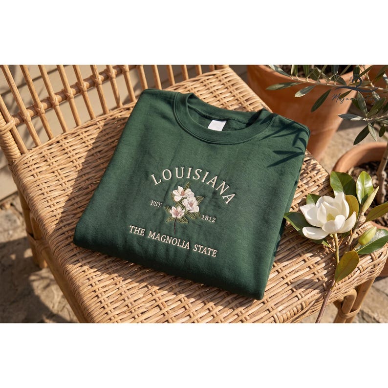May include: A dark green sweatshirt featuring the embroidered word "LOUISIANA", a floral design, and the text "THE MAGNOLIA STATE". The sweatshirt is placed on a wicker chair.