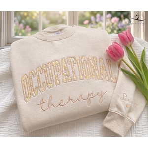 May include: A cream-colored crewneck sweatshirt featuring the text OCCUPATIONAL in yellow floral block letters and therapy in cursive script. The sleeve is embroidered with the name Sarah and a small heart. Two pink tulips rest on the fabric.