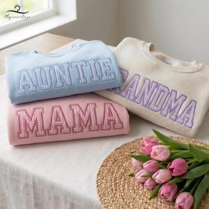May include: Three sweatshirts in pastel colors: light blue, pink, and cream. Each sweatshirt has a different word embroidered in a gingham pattern: "AUNTIE", "MAMA", and "GRANDMA". A bouquet of pink tulips rests on a woven mat.