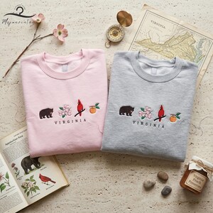May include: Two crewneck sweatshirts, one pink and one grey, embroidered with a bear, flowers, a cardinal, and a peach. The word "VIRGINIA" is embroidered below the design. The sweatshirts are part of a clothing store's collection.