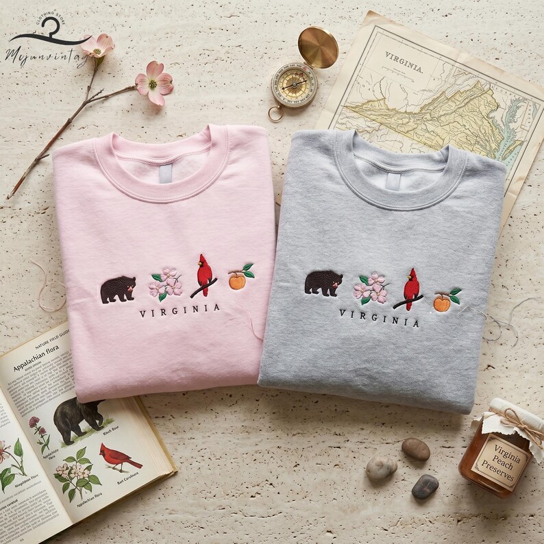 May include: Two crewneck sweatshirts, one pink and one grey, embroidered with a bear, flowers, a cardinal, and a peach. The word "VIRGINIA" is embroidered below the design. The sweatshirts are part of a clothing store's collection.