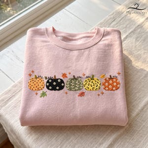 May include: A light pink crewneck sweatshirt featuring an embroidered row of five pumpkins across the chest. The pumpkins display various patterns including polka dots, leopard print, and floral motifs, accented by small embroidered leaves and stars.
