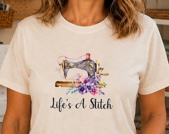 Könnte beinhalten: Ein cremefarbenes Kurzarm-T-Shirt mit einer Grafik einer Vintage-Nähmaschine, die mit violetten und blauen Blumen verziert ist. Unter der Illustration steht der Text Life's A Stitch in einer schwarzen Schreib- und Serifenschrift.