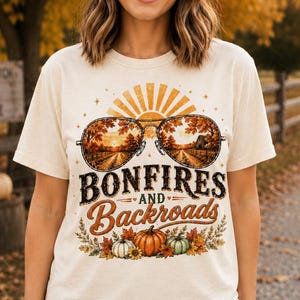 May include: A cream-coloured short-sleeve t-shirt featuring a graphic of aviator sunglasses reflecting a fall landscape with trees and a barn. Below the glasses, the text reads Bonfires and Backroads, accompanied by illustrations of pumpkins.