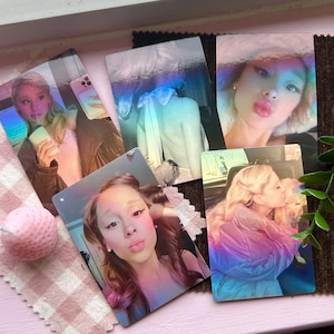 Puede incluir: Una colección de tarjetas fotográficas brillantes que muestran a una mujer en varias poses, con una tela de cuadros rosa y una vela rosa esférica. Las tarjetas tienen un efecto holográfico, con colores pastel suaves. El tema general es un producto temático de celebridades.
