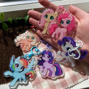 Op de afbeelding: Een verzameling kleurrijke, glinsterende sleutelhangers met ponythema. Elke sleutelhanger heeft een ander cartoon pony-personage met levendige kleuren, waaronder roze, geel, blauw en paars. De sleutelhangers hebben metalen sluitingen.