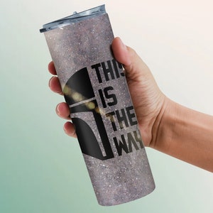 Pode incluir: Um copo de a&ccedil;o inoxid&aacute;vel com acabamento prateado brilhante e um gr&aacute;fico preto de um capacete mandaloriano com o texto "This is the way".