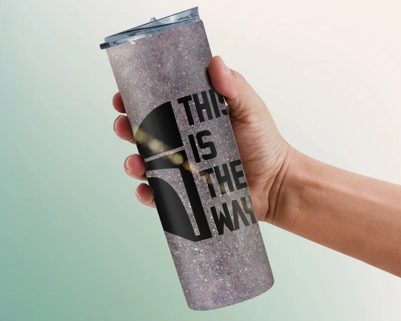 Pode incluir: Um copo de a&ccedil;o inoxid&aacute;vel com acabamento prateado brilhante e um gr&aacute;fico preto de um capacete mandaloriano com o texto "This is the way".