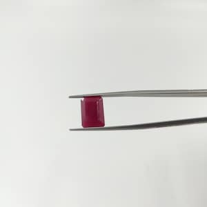 May include: A deep red rectangular gemstone held by tweezers against a white background.