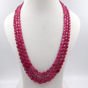 Può includere: Una collana a più fili composta da tre strati di perline lisce di forma ovale, di un colore rosso rosato intenso. Le perline sono uniformi per dimensioni e forma, infilate insieme per creare un effetto a strati che scende sul busto.