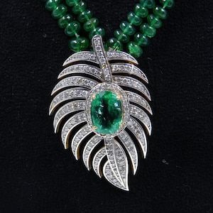 Op de afbeelding: Een edelstenen ketting met een blad-vormige hanger met een groot ovaal smaragdgroen midden, omringd door diamanten. De hanger hangt aan een streng kleine, ronde groene kralen. De achtergrond is zwart.
