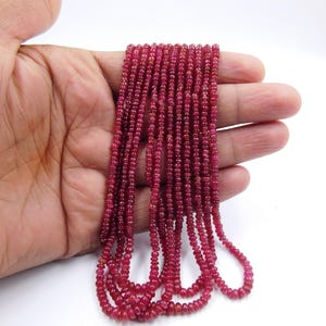 Puede incluir: Múltiples hebras de pequeñas cuentas redondas de color rojo intenso. Las cuentas están ensartadas, formando múltiples hebras. Las cuentas son de un color rojo oscuro intenso y parecen pulidas. Las cuentas se sostienen en una mano sobre un fondo blanco.