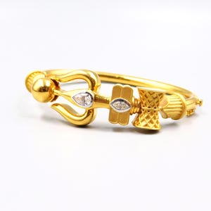 Op de afbeelding: Een goudkleurige armband met ingewikkelde details. De armband heeft een drietandontwerp met een druppelvormige edelsteen, een rechthoekig element met een edelsteen en een decoratief element. De armband ligt op een witte achtergrond.