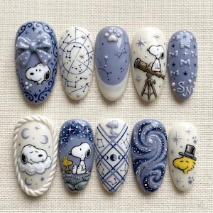 May include: A set of ten oval-shaped, painted fingernails. Each nail features a different design, including Snoopy, constellations, bows, and paw prints, in shades of blue, white, and yellow. The nails are decorated with intricate details and patterns.